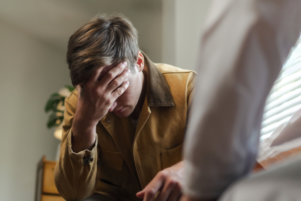 A man in a brown jacket sits with his head in his hand, appearing distressed.