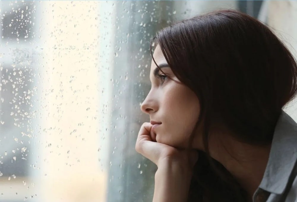 A woman gazing out of a window on a rainy day, her face resting on her hand.